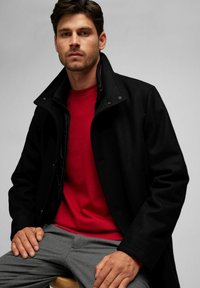 Schwarzer Wollmischmantel mit hohem Kragen, doppelter Knopfverschluss und dezentem Futter, getragen über einem roten Rundhalsausschnitt-Pullover und grauen Hosen.