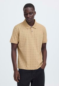 Polo beige en tissu texturé avec petits motifs noirs, col à revers et patte de boutonnage deux boutons ; associé à un pantalon foncé.