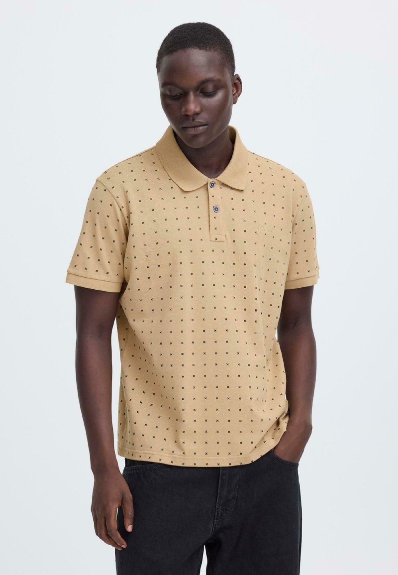 Polo beige en tissu texturé avec petits motifs noirs, col à revers et patte de boutonnage deux boutons ; associé à un pantalon foncé.