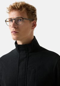 Młody mężczyzna z kręconymi, blond włosami i okrągłymi okularami, ubrany w czarną kurtkę z wysokim kołnierzem, patrzy nieco w lewo na jednolite, jasne tło.