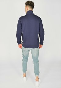 Tummansininen neule, jossa on korkea kaulus ja sileä tekstuuri. Yhdistetty vaaleansinihin kapeisiin farkkuihin ja valkoisiin lenkkareihin.