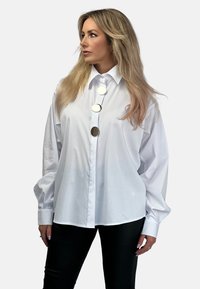 Biała, ponadwymiarowa koszula z guzikami, z zaostrzonym kołnierzykiem, z trzema dużymi srebrnymi guzikami z przodu i długimi, puszystymi rękawami.