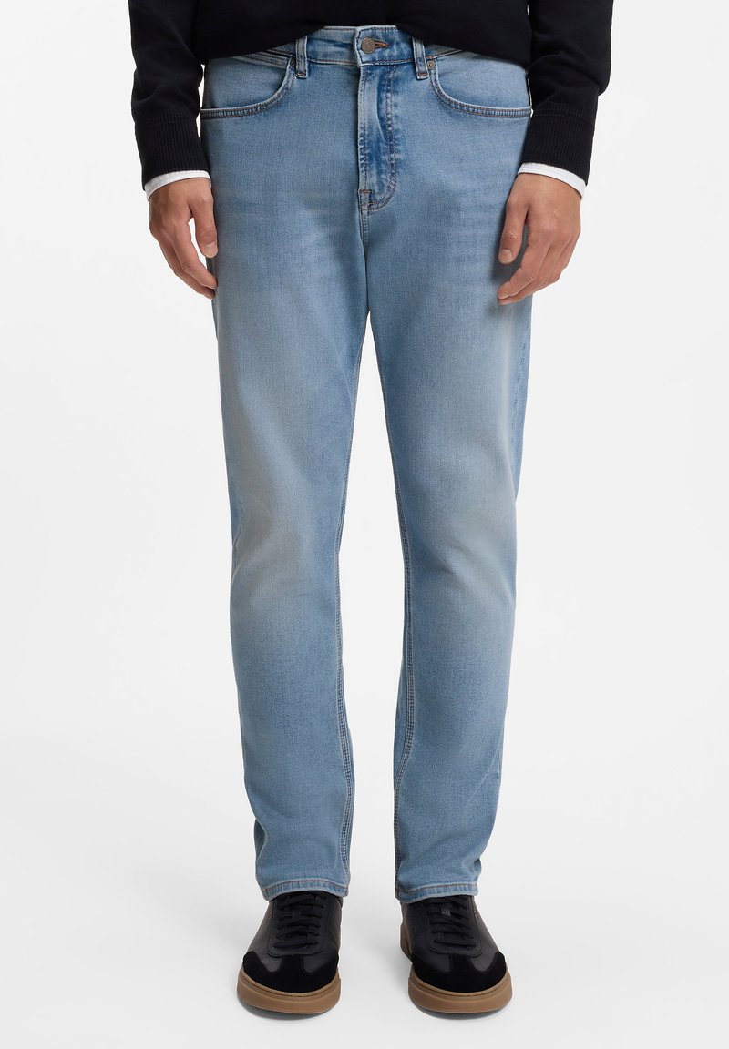 Svetlomodré denimové rifle s priamym strihom, viditeľným prešívaním a prednými vreckami. Zladené s čiernymi teniskami na jednoduchom pozadí.