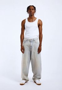 Světle modré džínové kalhoty s širokými nohavicemi a vybledlými detaily, zkombinované s bílým tílkem a bílými teniskami, stojící před jednobarevným pozadím.