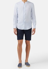 Jasnoniebieska i biała w paski koszula z długim rękawem, zapinana na guziki, z kołnierzem typu band, w połączeniu z granatowymi szortami i niebieskimi espadrylami.