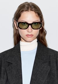 Černé obdélníkové sluneční brýle se zelenými skly, nošené s světle modrou mikinou a bílým rolákem pod tmavým strukturovaným blejzrem.