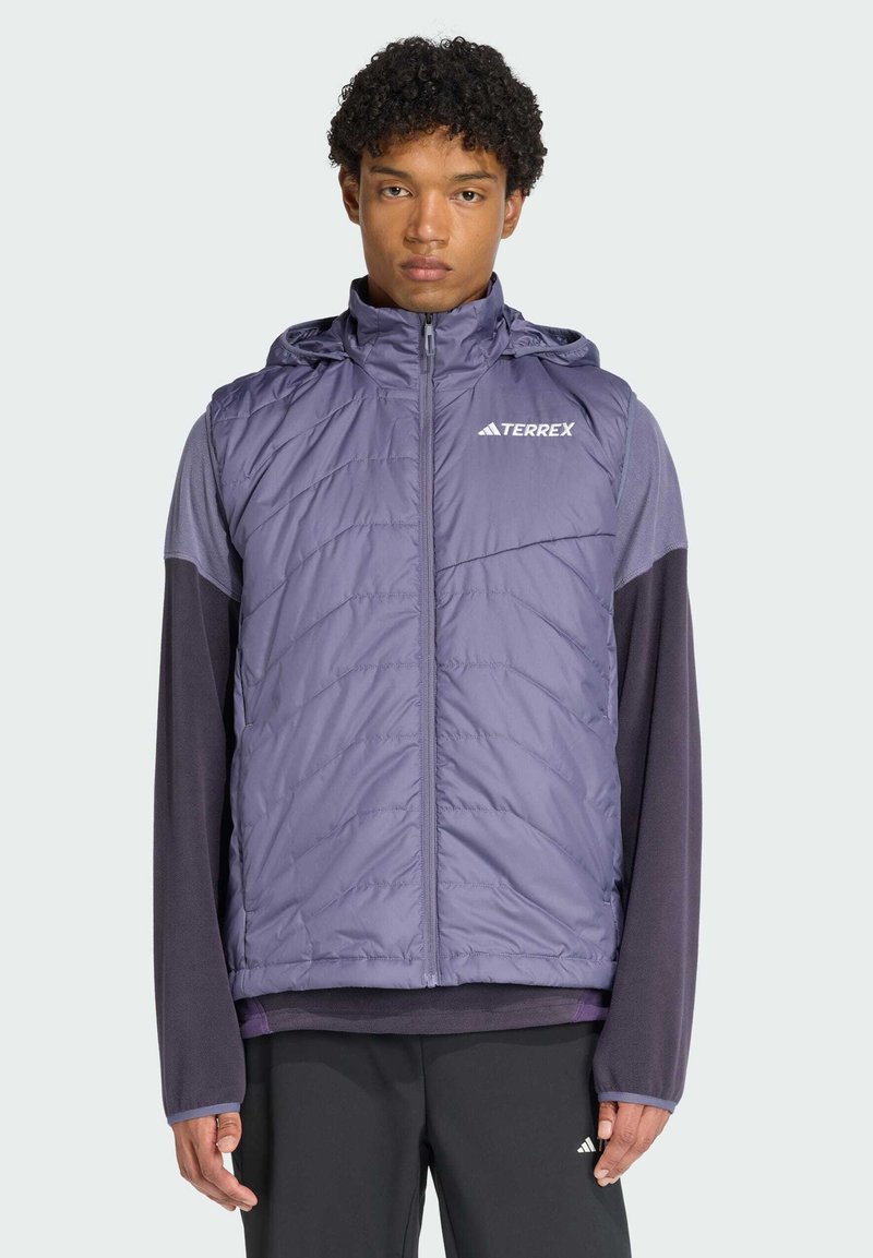 Jeune homme portant un gilet matelassé sans manches violet avec une capuche par-dessus une chemise à manches longues et un pantalon noir, debout devant un fond uni.