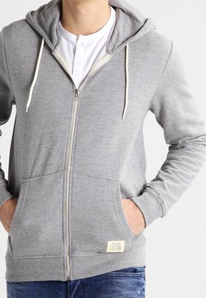 Hombre con una sudadera con cremallera de color gris claro, con cordones y bolsillos frontales, con las manos dentro de los bolsillos sobre unos jeans azules.