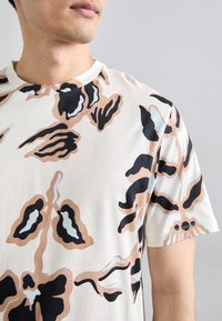 T-shirt a maniche corte in base crema con un motivo astratto audace in nero, marrone e azzurro chiaro. Presenta un colletto rotondo e una texture del tessuto.