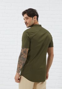 Short-sleeve, olive green shirt with a mandarin collar. Smooth texture, slim fit design, and slightly curved hem. Model displays intricate tattoos.