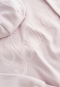 Rozā kapuce ar izšūtu tekstu "Baker" un "BY TED BAKER." Tam ir mīksta auduma tekstūra un regulējama kapuce.