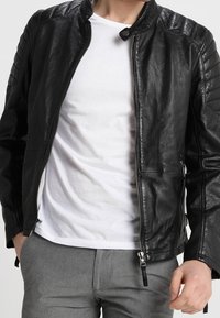 Giacca da motociclista in pelle nera con collo alla coreana, dettagli a costine sulle spalle e cerniere argentate. Indossata sopra una camicia bianca.