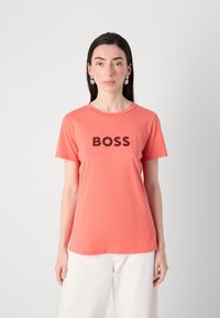 Modelis valkā korallkrāsas t-kreklu ar uzrakstu "BOSS" melnās burtu izdrukā, apvienojot to ar baltām biksēm, uz vienkrāsaina pelēka fona.