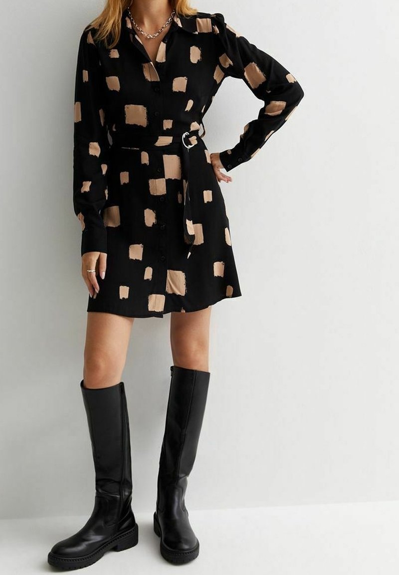 Femme portant une robe-chemise noire avec des motifs carrés beige, taille ceinturée, et bottes noires montant jusqu'aux genoux, debout devant un mur blanc uni.