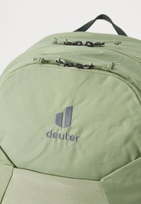 Grüner Stoff-Rucksack mit glatter Textur, doppelten Reißverschlussfächern und einem grauen Logo, das prominent auf der Vorderseite angezeigt wird.