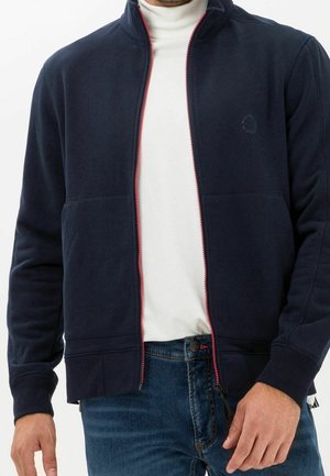 Man draagt een marineblauwe jas met rits en rode rand over een witte coltrui en blauwe spijkerbroek, staat met ontspannen handen.