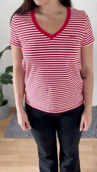 Woman wearing a red and white striped V-neck T-shirt and black pants, standing indoors with plants and a gray rug in the background.