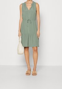 Abito senza maniche di un verde chiaro con scollatura a V, spalle drappeggiate, vita con fiocco e gonna svasata. Abbinato a una borsa crema e sandali nudi.