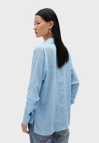 Camicia a maniche lunghe blu chiaro con patta bottonata, motivo testurizzato e spacchi laterali. Il tessuto appare morbido e traspirante.