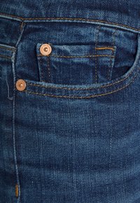 Džínové kalhoty v tmavě modré barvě s kontrastním žlutým šitím, dvěma měděnými nýty a malým detailem přední kapsy.