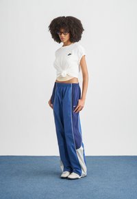 Λευκό βαμβακερό T-shirt με κόμπο στο μπροστινό μέρος, συνδυασμένο με μπλε παντελόνι φόρμας με λευκές πλευρικές ρίγες και μικρό λογότυπο. Υφή υφάσματος.