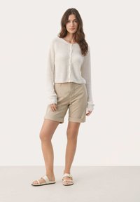 Shorts de algodón beige y un cárdigan blanco de canalé con botones. La modelo lleva sandalias de color claro, mostrando un atuendo casual de verano.