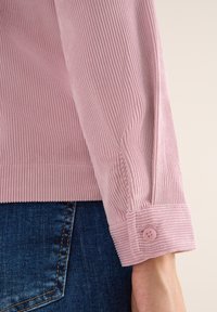 Puño de camisa de cortón rosa con estrías verticales, que presenta un cierre de botón y detalles de costura. Combinado con denim azul.