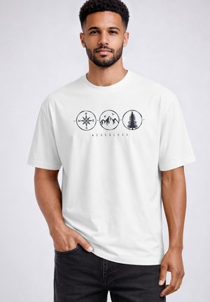 Mann, der ein weißes T-Shirt mit schwarzen Kompass-, Berg- und Baum-Symbolen über dem Wort „NEVERTHELESS“ trägt, steht mit einer Hand in der Tasche.
