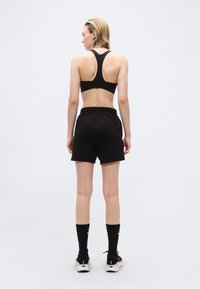 Γυναίκα με κοντά ξανθά μαλλιά που φοράει μαύρο αθλητικό σουτιέν τύπου racerback, μαύρο σορτς, μαύρες κάλτσες μέχρι τη μέση της γάμπας και μαύρα αθλητικά παπούτσια, στέκεται γυρισμένη με την πλάτη.