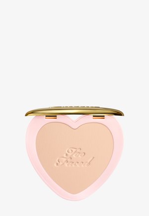 Poudre compacte en forme de cœur avec poudre beige embossée "Too Faced", boîtier rose et couvercle doré brillant ouvert sur fond blanc.
