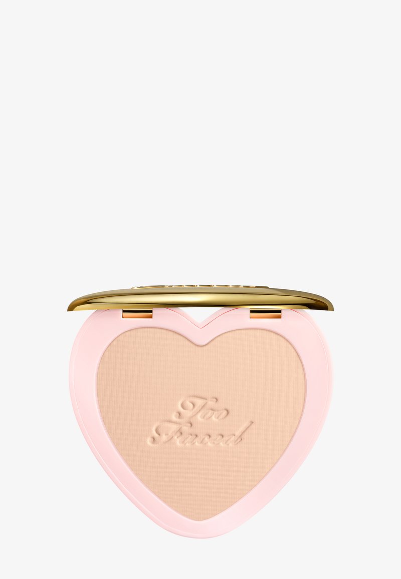 Poudre compacte en forme de cœur avec poudre beige embossée "Too Faced", boîtier rose et couvercle doré brillant ouvert sur fond blanc.