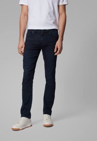Man som bär mörkblå slim-fit jeans, vit t-shirt och vita sneakers med bruna sulor ståendes mot en grå bakgrund.