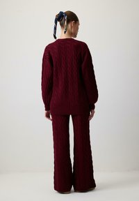 Sieviete, ģērbta atbilstošā bordo krāsas adītā svīterī un platā stila biksēs, stāv ar muguru pret kameru, pret vienkāršu gaišu sienu.