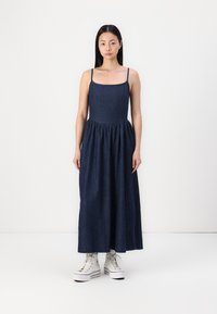 Jeans haljina maxi dužine s tankim naramenicama, prilagođenim tijelom i skupljenom suknjom. Tamnoplava, glatka tkanina s blagim sjajem, u kombinaciji s bijelim tenisicama.