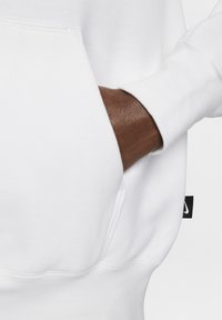 Witte trui met een kangoeroezak, zachte textuur en een zijkantlabel met een logo. De mouw onthult gedeeltelijk een donkerbruine hand.