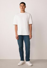 Fehér, rövid ujjú póló, laza szabású, sötétkék farmerral és fehér sportcipővel párosítva. Egyszerű dizájn, hétköznapi stílus.