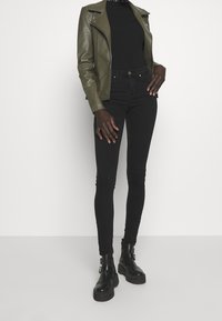 Mujer vestida con una chaqueta de cuero verde oliva, un suéter de cuello alto negro, jeans ajustados negros y botas de tobillo negras con hebilla, de pie contra un fondo blanco.