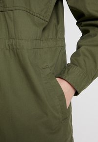 Olive green jacket with a smooth, woven texture, featuring a chest pocket and a hand pocket on the side for practicality.