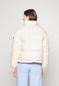 Crème-kleurige pufferjas met een hoge kraag, een geborduurd ontwerp en elastische manchetten. G gedragen over lichtblauwe broeken.