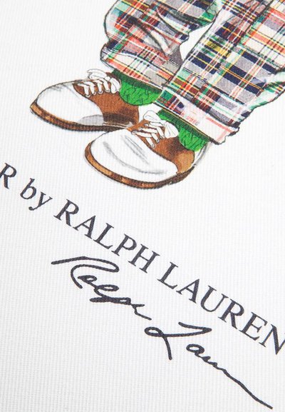 Παιδικά πόδια με καφέ και λευκά παπούτσια, πράσινες κάλτσες, καρό παντελόνι, πάνω από το κείμενο "R by Ralph Lauren" και την υπογραφή σε λευκό ύφασμα.