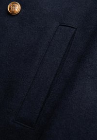 Marineblauer Wollstoff mit strukturierter Oberfläche, ausgestattet mit einer rechteckigen Tasche und einem goldenen Knopfdetail, das ein sauberes und maßgeschneidertes Design zeigt.
