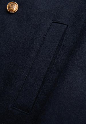 Tessuto di lana blu navy con finitura texturizzata, caratterizzato da una tasca rettangolare e un dettaglio con bottone dorato, che mette in mostra un design pulito e sartoriale.