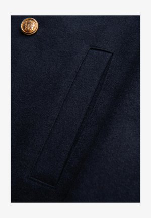 Tessuto di lana blu navy con finitura texturizzata, caratterizzato da una tasca rettangolare e un dettaglio con bottone dorato, che mette in mostra un design pulito e sartoriale.