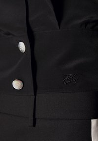 Černá tkanina s hladkým povrchem má velké, kulaté, lesklé knoflíky a vyšívaný logo detail. Design zahrnuje strukturované panely.