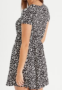 Femme portant une robe noire à manches courtes avec un motif floral blanc, cintrée à la taille, vue de dos et de côté.