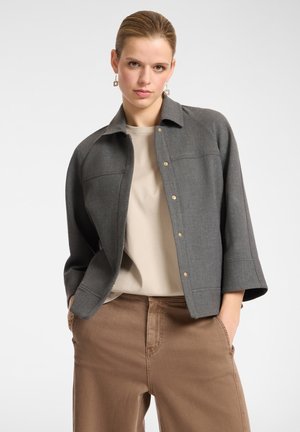 Chaqueta estructurada gris con cuello tipo solapa, que presenta botones de presión dorados y mangas de tres cuartos, combinada con una blusa beige y pantalones marrones.