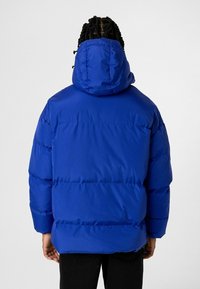 Blauwe, gewatteerde winterjas met een capuchon; heeft horizontale naden en een gladde, glanzende afwerking. Draagt een zwarte broek.