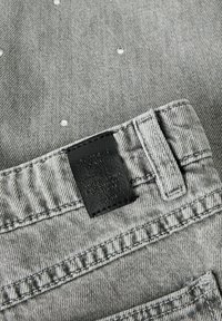 Hallide ninat kootud hallid teksased, millel on tekstuuriga viimistlus, must nahast silt ja dekoratiivsed hõbedased nupud kanga peal.