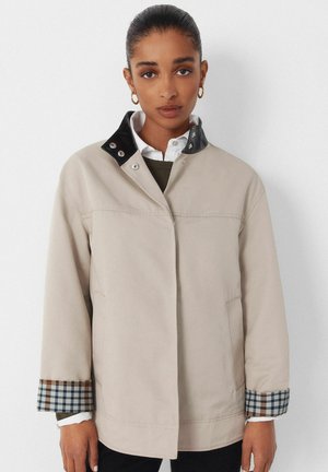 Frau trägt eine beige Jacke mit schwarzem Kragen und kariertem Manschettenfutter, weißes Hemd und goldene Creolen vor einfarbigem Hintergrund.