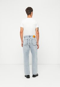 Lyseblå denimjeans med en avslappet passform, med et gult merke på baklommen og subtil falming gjennom hele. Svarte sko er synlige.